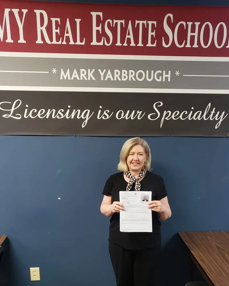 A student holding her passing exam score in front of the MY Real Estate School banner